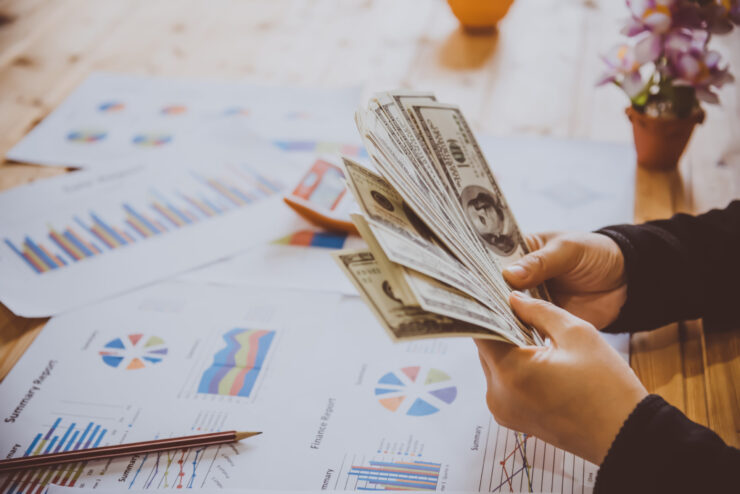 Person counting a stack of US dollar bills at a desk with financial charts, graphs, and reports spread out.