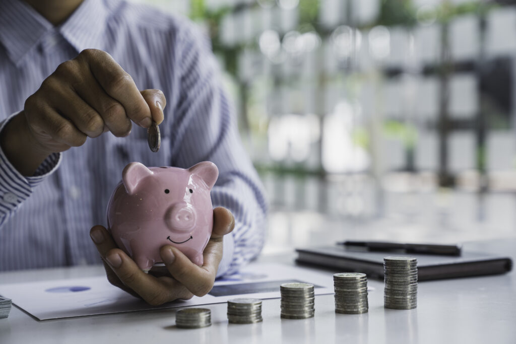 A person in a striped shirt places a coin into a pink piggy bank, with stacks of coins on a table. The scene conveys savings and financial growth.