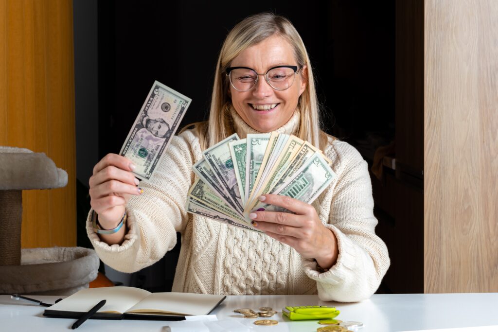 Smiling woman wearing glasses and a sweater holds a fan of U.S. dollar bills at a desk with coins and a notebook, conveying a sense of joy and financial success.