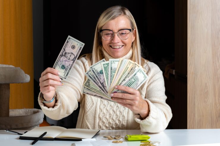 Smiling woman wearing glasses and a sweater holds a fan of U.S. dollar bills at a desk with coins and a notebook, conveying a sense of joy and financial success.