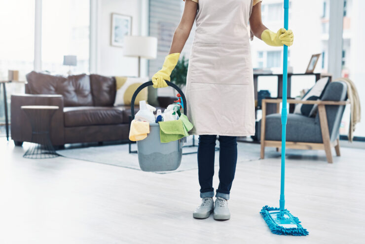 Person wearing a white apron and yellow rubber gloves cleaning a modern living room, holding a mop and a bucket filled with cleaning supplies including spray bottles and microfiber cloths. Bright, tidy home interior with a sofa, armchair, and large windows in the background. Professional house cleaning, domestic chores, sanitation, and housekeeping concept with focus on tools and cleanliness.