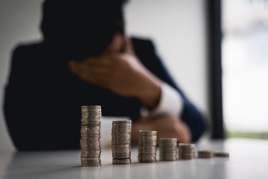 A man in a suit is sitting at a table with stacks of coins. He rests his head on his hand, appearing stressed. The focus is on the coins.