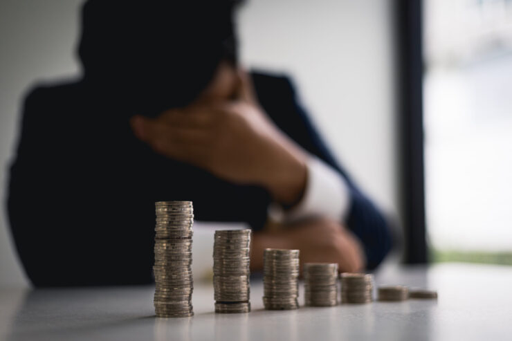 A man in a suit is sitting at a table with stacks of coins. He rests his head on his hand, appearing stressed. The focus is on the coins.