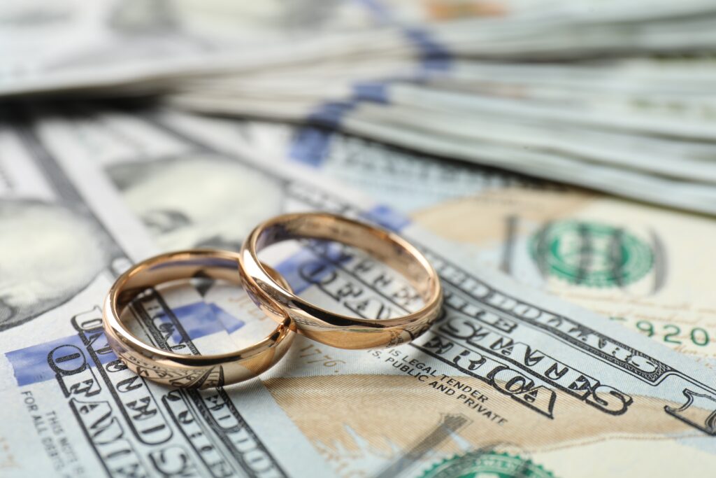 Two gold wedding rings rest on a pile of hundred-dollar bills, symbolizing the financial aspects of marriage. The tone suggests wealth and commitment.