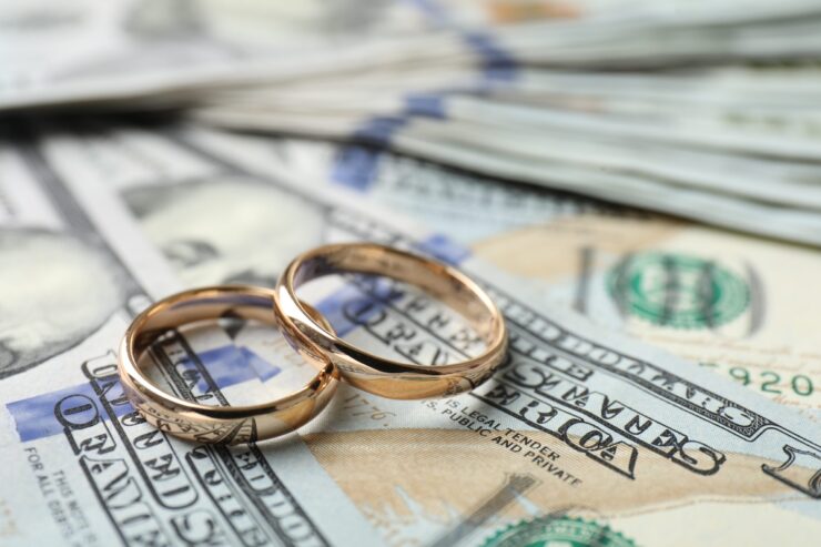 Two gold wedding rings rest on a pile of hundred-dollar bills, symbolizing the financial aspects of marriage. The tone suggests wealth and commitment.