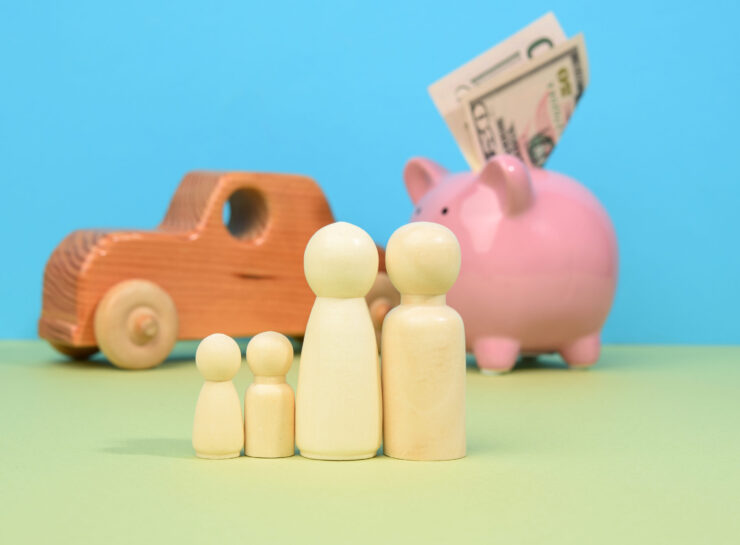 Wooden family figurines stand on a green surface, with a pink piggy bank holding dollar bills and a wooden toy car in the background, conveying financial planning.