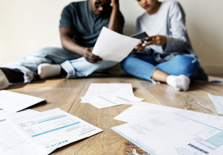 Couple sitting on wooden floor reviewing bills and invoices, stressed about finances, holding credit card and paperwork, home budgeting, debt management concept, scattered financial documents, indoor lifestyle scene