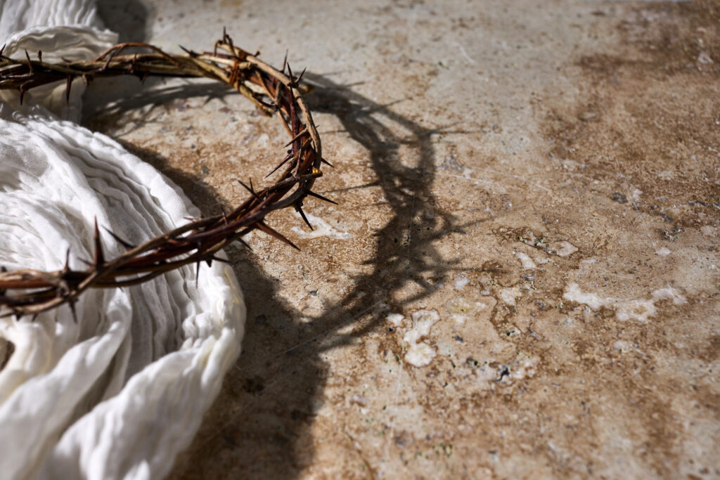 A crown of thorns rests on a stone surface beside a folded, white cloth. Shadows are cast on the ground, evoking a solemn and contemplative mood.