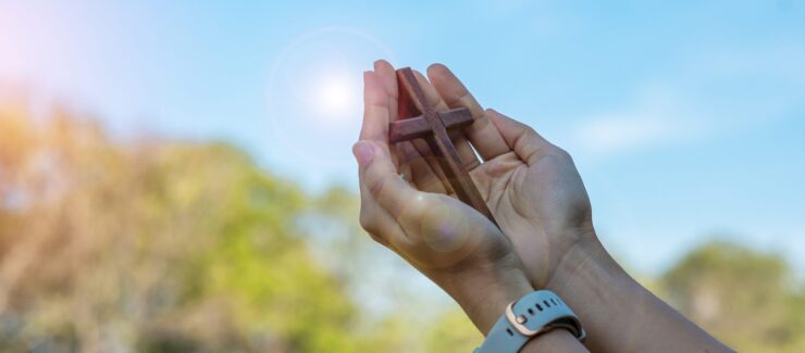 Hands holding wooden Christian cross toward sunlight, faith and spirituality concept, prayer gesture outdoors, bright sky with lens flare, religious symbol of hope and belief, close-up devotional scene, natural background
