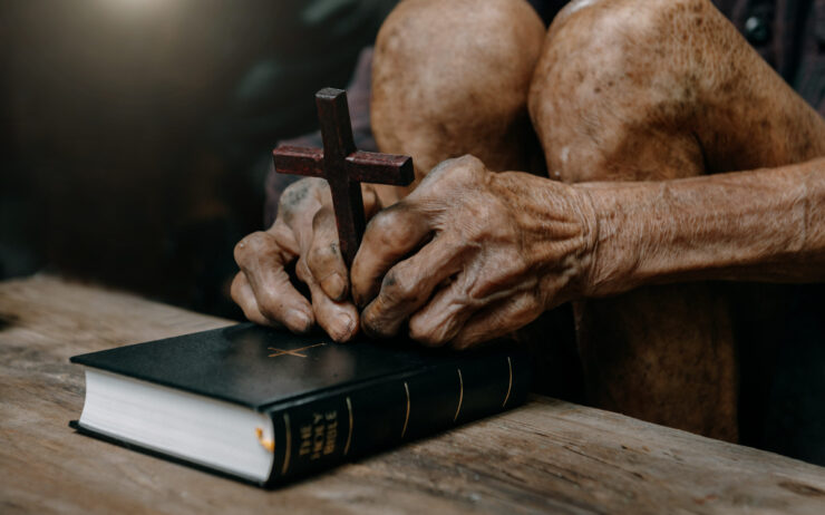 Elderly hands clasp a wooden cross atop a closed Bible on a wooden table, conveying faith and devotion, with warm, gentle lighting.