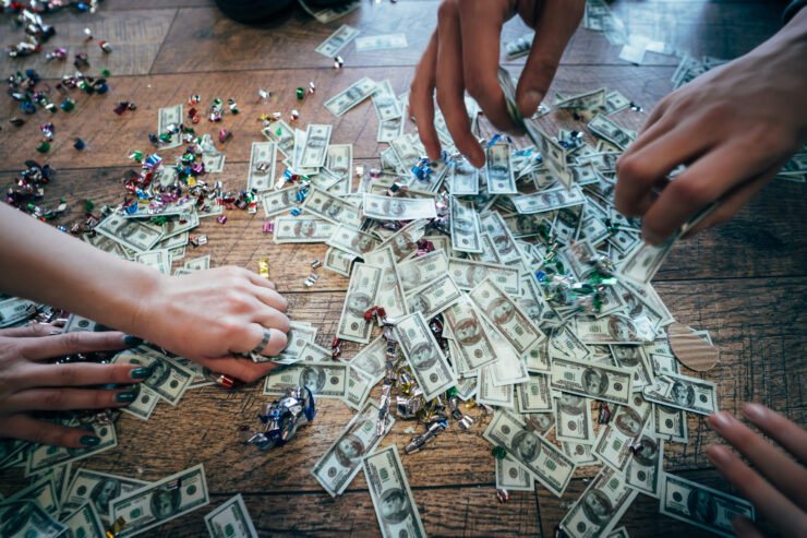 Hands reaching for scattered dollar bills and colorful candy wrappers on a wooden floor, conveying excitement and urgency.
