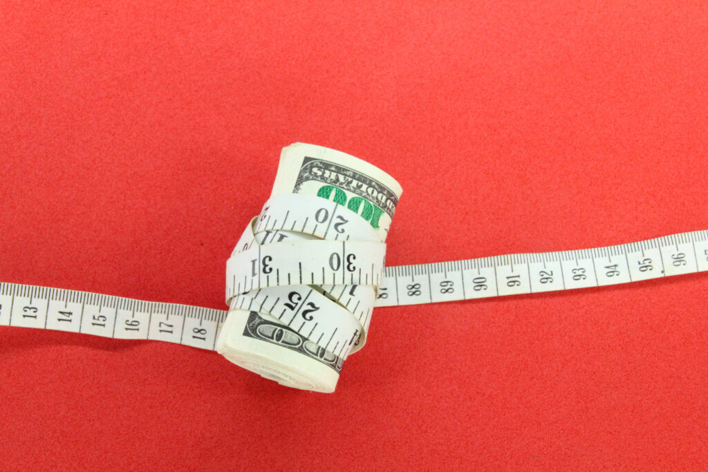 A roll of U.S. dollar bills wrapped tightly by a measuring tape on a red background, symbolizing financial constraint and budgeting.
