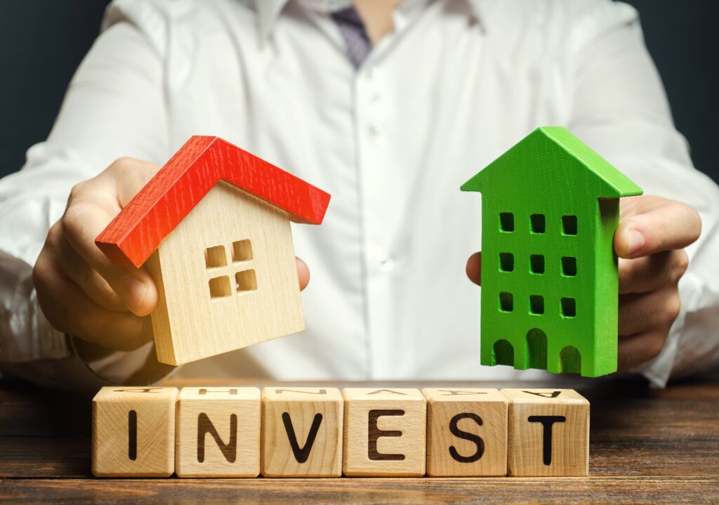 Person holding wooden models of a house and apartment building over lettered blocks spelling "INVEST," suggesting real estate investment choices.