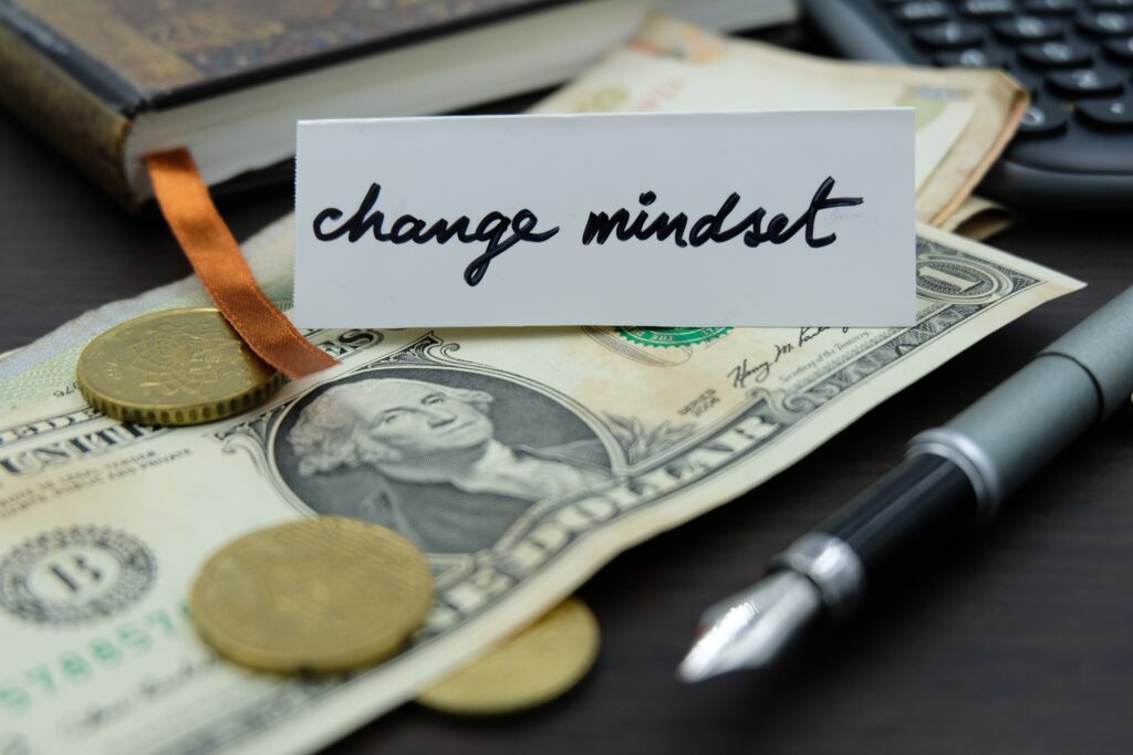 A card with "change mindset" rests on dollar bills and coins, surrounded by a pen, calculator, and closed book, conveying finance and transformation themes.