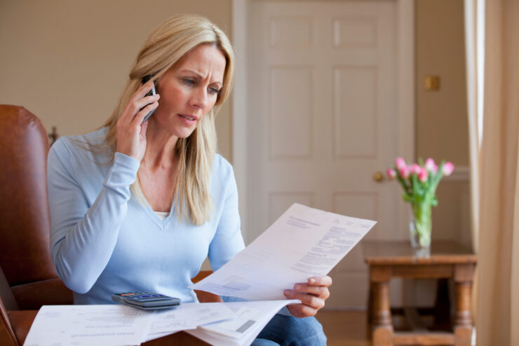 A woman with long blonde hair looks concerned as she reviews bills and talks on a cellphone. Papers, a calculator, and a vase of tulips are on the table.