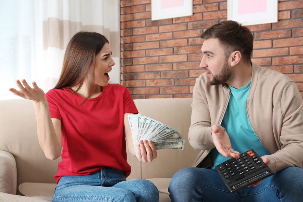 A woman in a red shirt, holding money, expresses surprise while a man in a beige jacket, holding a calculator, appears frustrated on a sofa.