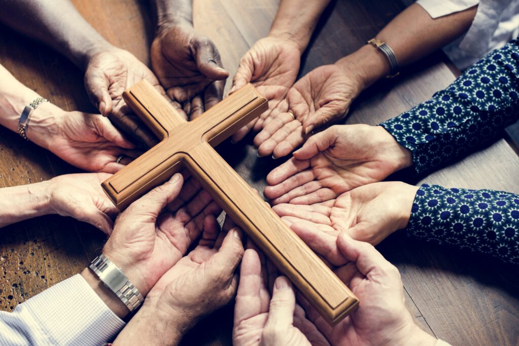 A group of diverse hands gathers, palms up, around a wooden cross on a table. The image conveys unity, faith, and communal support.