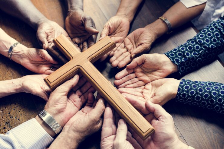 A group of diverse hands gathers, palms up, around a wooden cross on a table. The image conveys unity, faith, and communal support.