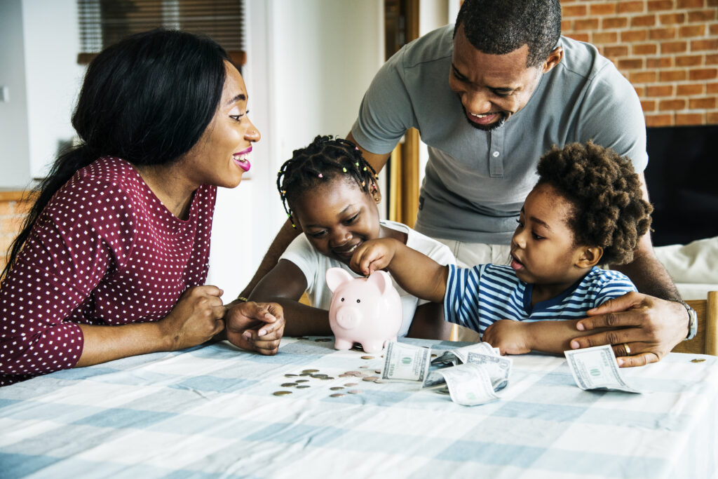 A joyful family gathers around a table with a pink piggy bank, coins, and bills. The parents and two children smile, sharing a moment of financial learning.