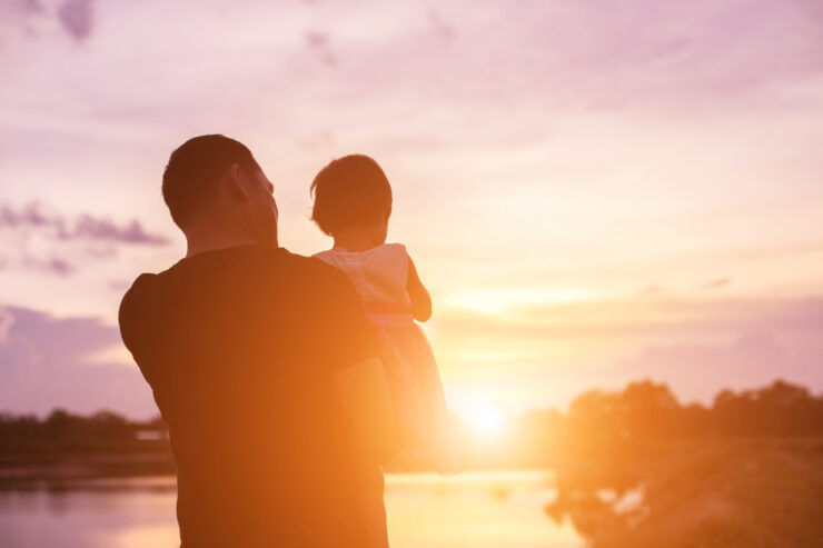 A man holds a young child, silhouetted against a vibrant sunset by a river. The scene conveys warmth and tranquility, with orange and purple hues.