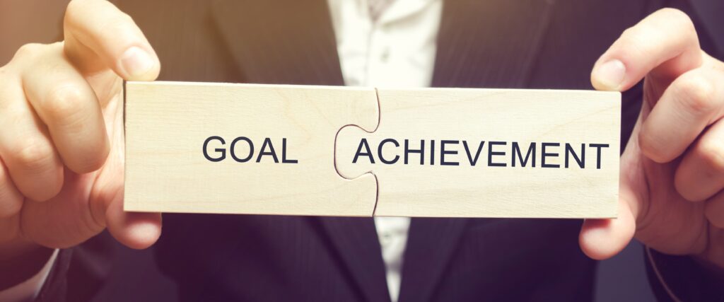 Two wooden puzzle pieces held by a person in a suit, fitting together to form the words "Goal" and "Achievement," symbolizing success and completion.