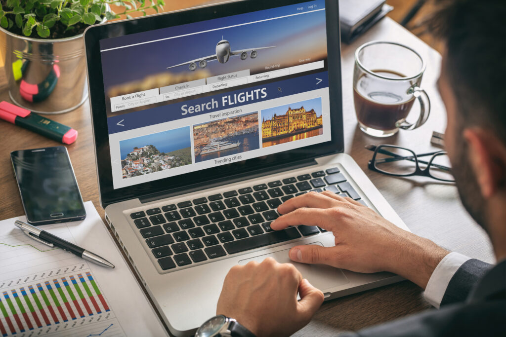 Man using a laptop showing a flight search webpage. Desk includes charts, planner, smartphone, glasses, pens, and a coffee cup, conveying productivity.
