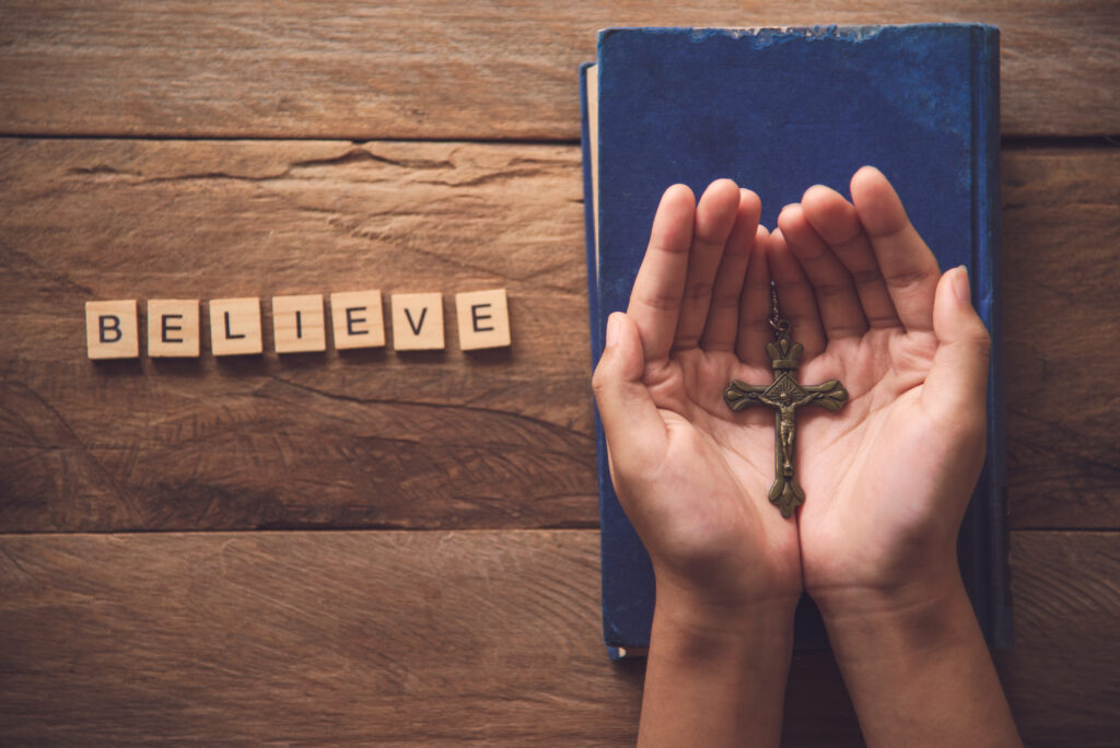 Hands cradle a metal cross over a blue book on a wooden surface. To the left, Scrabble tiles spell "BELIEVE," conveying faith and spirituality.