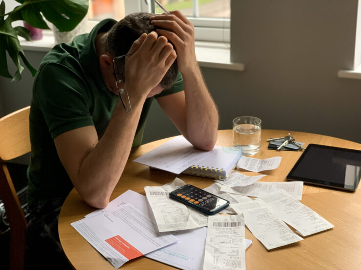 Man in green shirt sits at a wooden table overwhelmed with paperwork, calculator, bills, and receipts, conveying stress and financial pressure.
