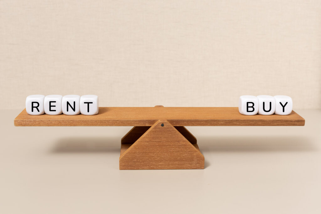 Wooden balance scale with "RENT" on left and "BUY" on right, symbolizing a choice between renting and buying. Neutral background, contemplative tone.