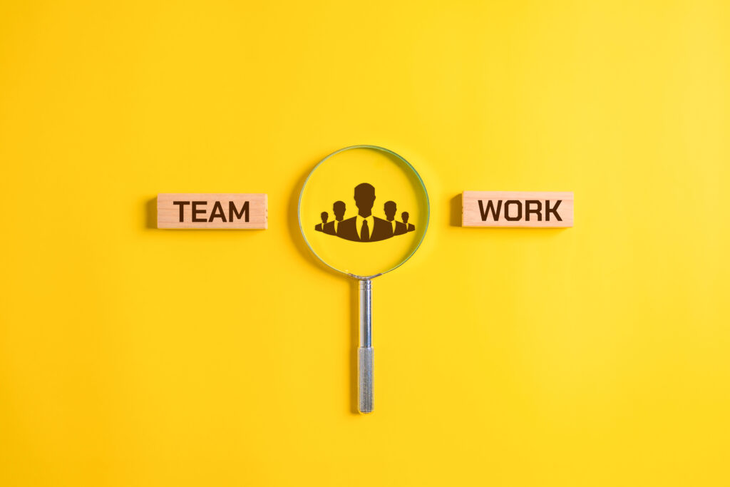 A magnifying glass highlights silhouettes of people, symbolizing teamwork, between wooden blocks labeled "TEAM" and "WORK" on a yellow background.
