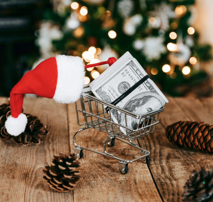A Santa hat draped over a small shopping cart filled with dollar bills on a wooden table, surrounded by pinecones, with a blurred, festive background.