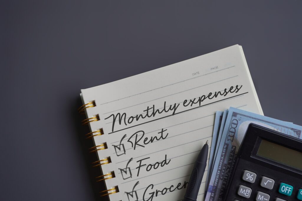 Open notebook with "Monthly expenses" listed, including rent, food, and groceries. Nearby are a pen, calculator, and a stack of dollar bills.