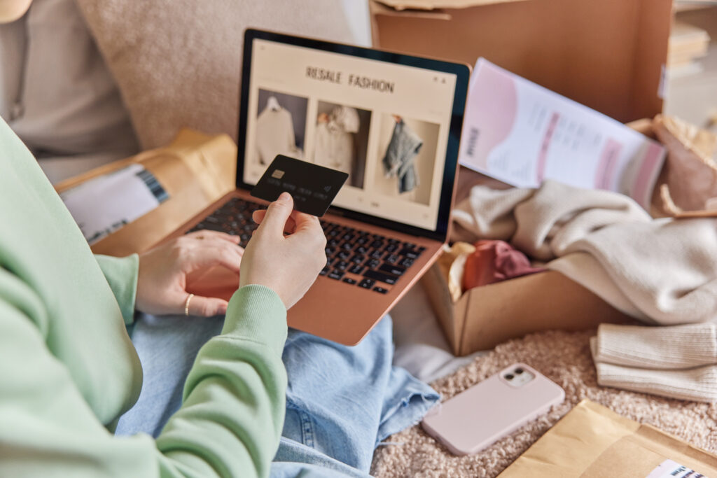 Person shopping for clothes online on a laptop while holding a credit card, surrounded by open cardboard shipping boxes, folded garments, and packaging materials on a cozy carpeted floor, depicting e-commerce fashion purchase, online retail, home delivery, and digital payment concept; laptop screen displays “RESALE FASHION” with clothing product images, smartphone lying nearby.