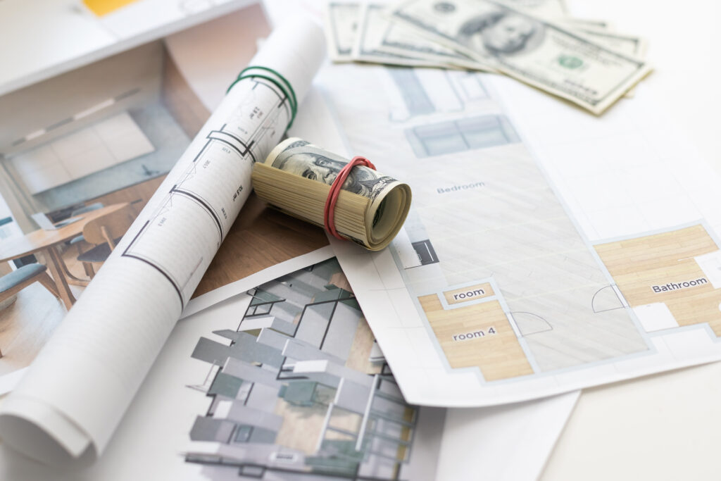 Rolled architectural plans, floor layouts, and a few U.S. dollar bills spread on a table, suggesting building design and budgeting.