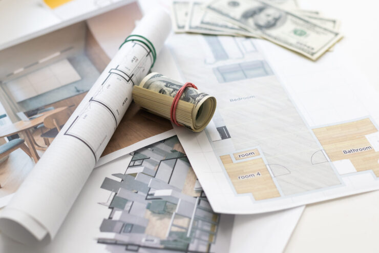 Rolled architectural plans, floor layouts, and a few U.S. dollar bills spread on a table, suggesting building design and budgeting.