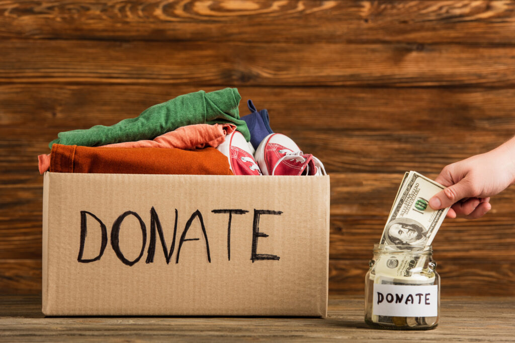 A cardboard box labeled "DONATE" contains folded clothes and shoes. Nearby, a hand places dollar bills into a donation jar, evoking a spirit of giving.