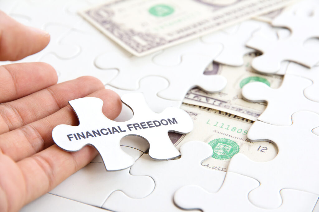 A hand holds a puzzle piece labeled "Financial Freedom" over incomplete pieces revealing dollar bills, symbolizing economic independence and achievement.