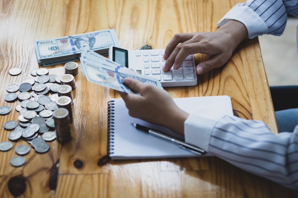 A person in a striped shirt counts dollar bills while using a calculator. Nearby, a stack of coins, a notebook, and a pen rest on a wooden table.