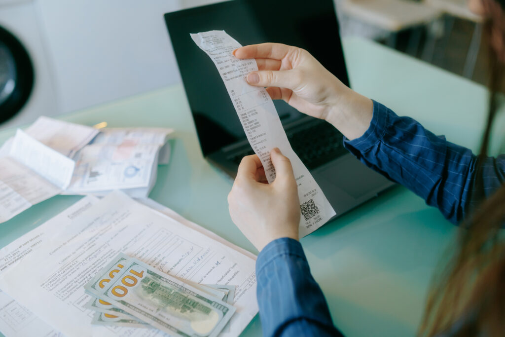 A person examines a detailed receipt while seated at a clean desk. Cash and documents are spread out, and a laptop is open, indicating financial organization.