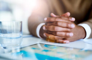 A person sitting at a desk with hands clasped in thought beside a glass of water and tablet, symbolizing careful planning and reflection while reviewing a monthly budget.