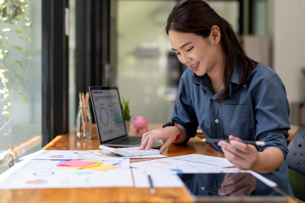 A woman in a blue shirt smiles while working at a desk with a laptop, calculator, and papers with charts. Sunlight streams through nearby windows.