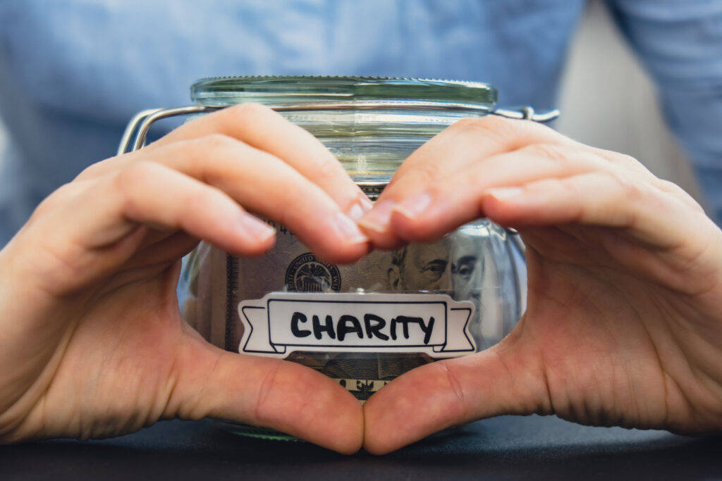 Female showing heart sign with hands Saving Money In Glass Jar filled with Dollars banknotes. CHARITY transcription in front of jar. Managing personal finances extra income for future insecurity background